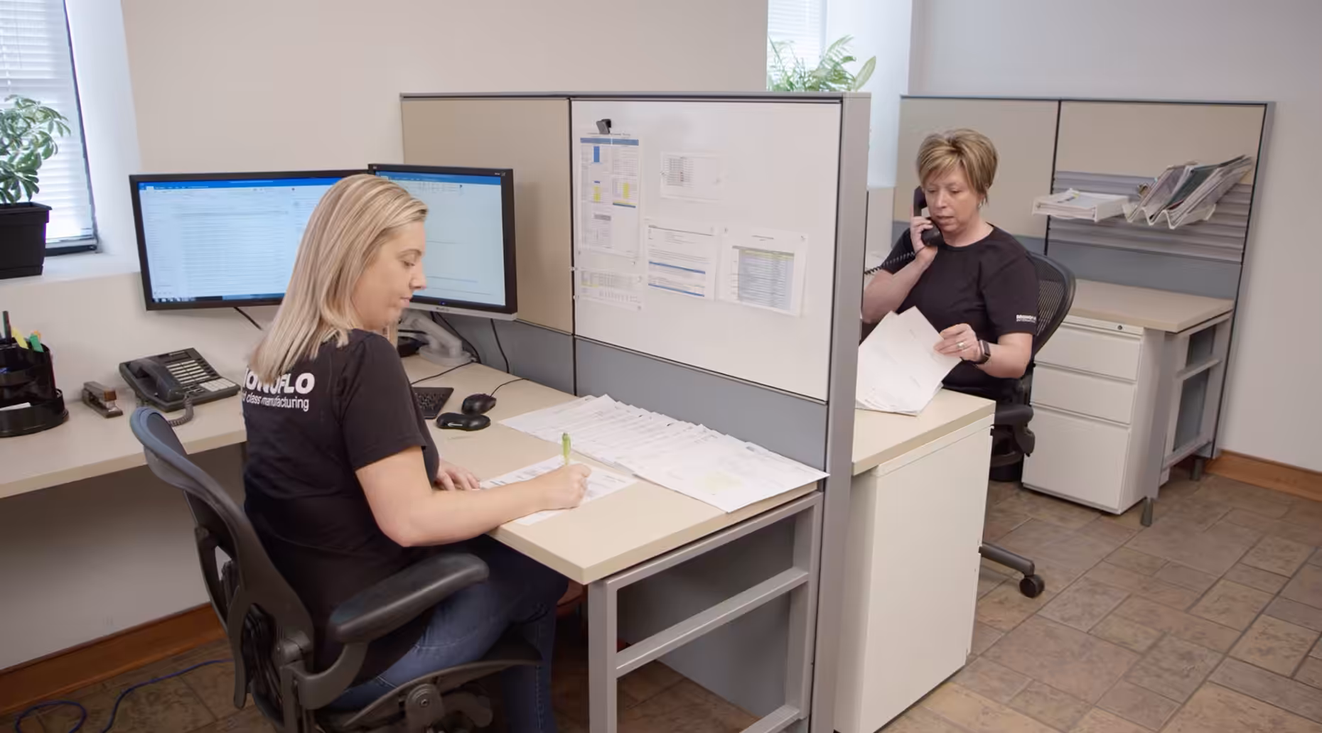 Two office workers in cubicles handling paperwork and phone calls at desks with computers in a modern workplace.