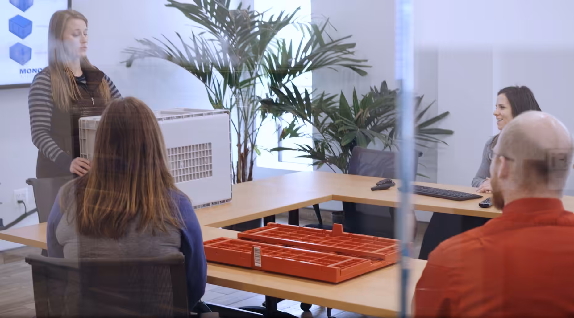 Team meeting where a presenter shows a large plastic container while others review red plastic parts on the table.