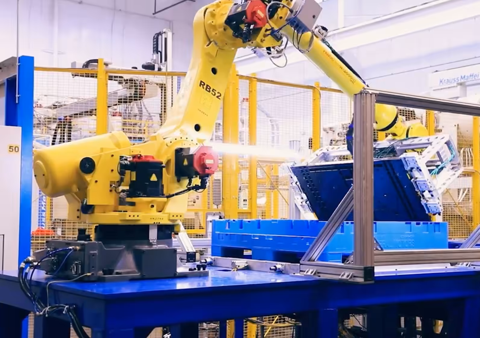 Automated robotic arm operating in a manufacturing facility with machinery and storage containers.