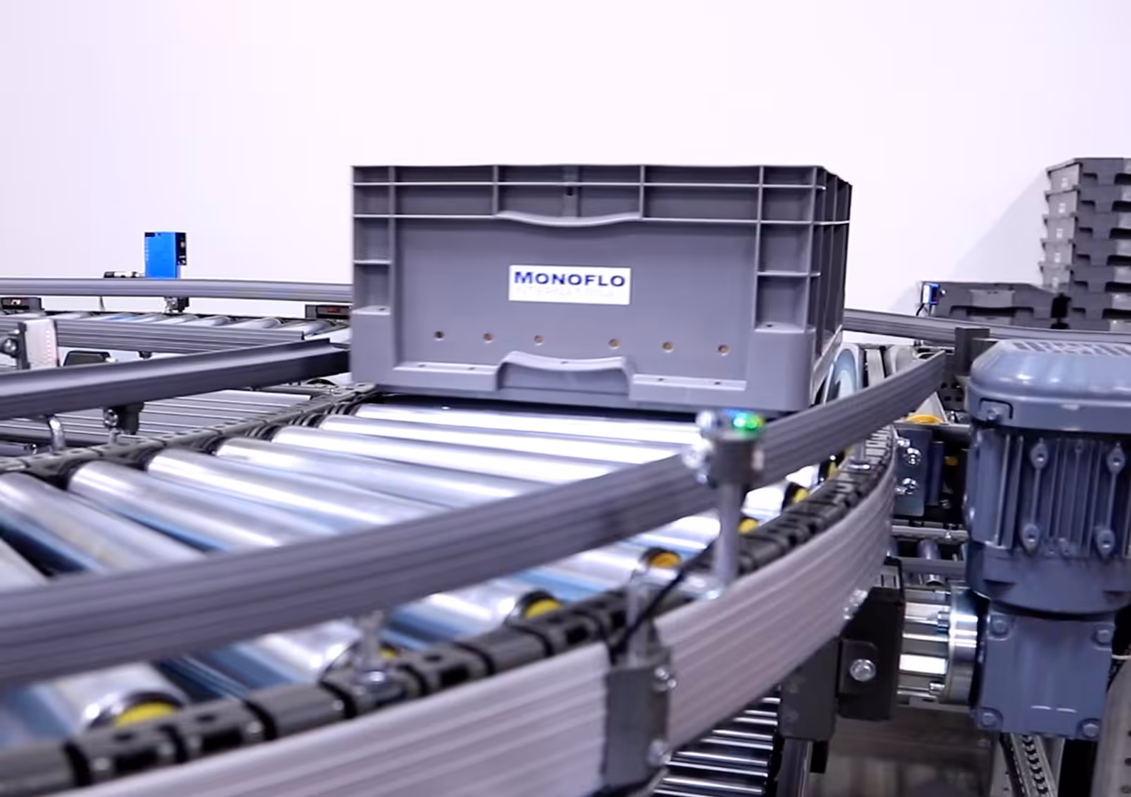 Gray plastic tote on an automated roller conveyor system in a warehouse, with motor and sensors visible.