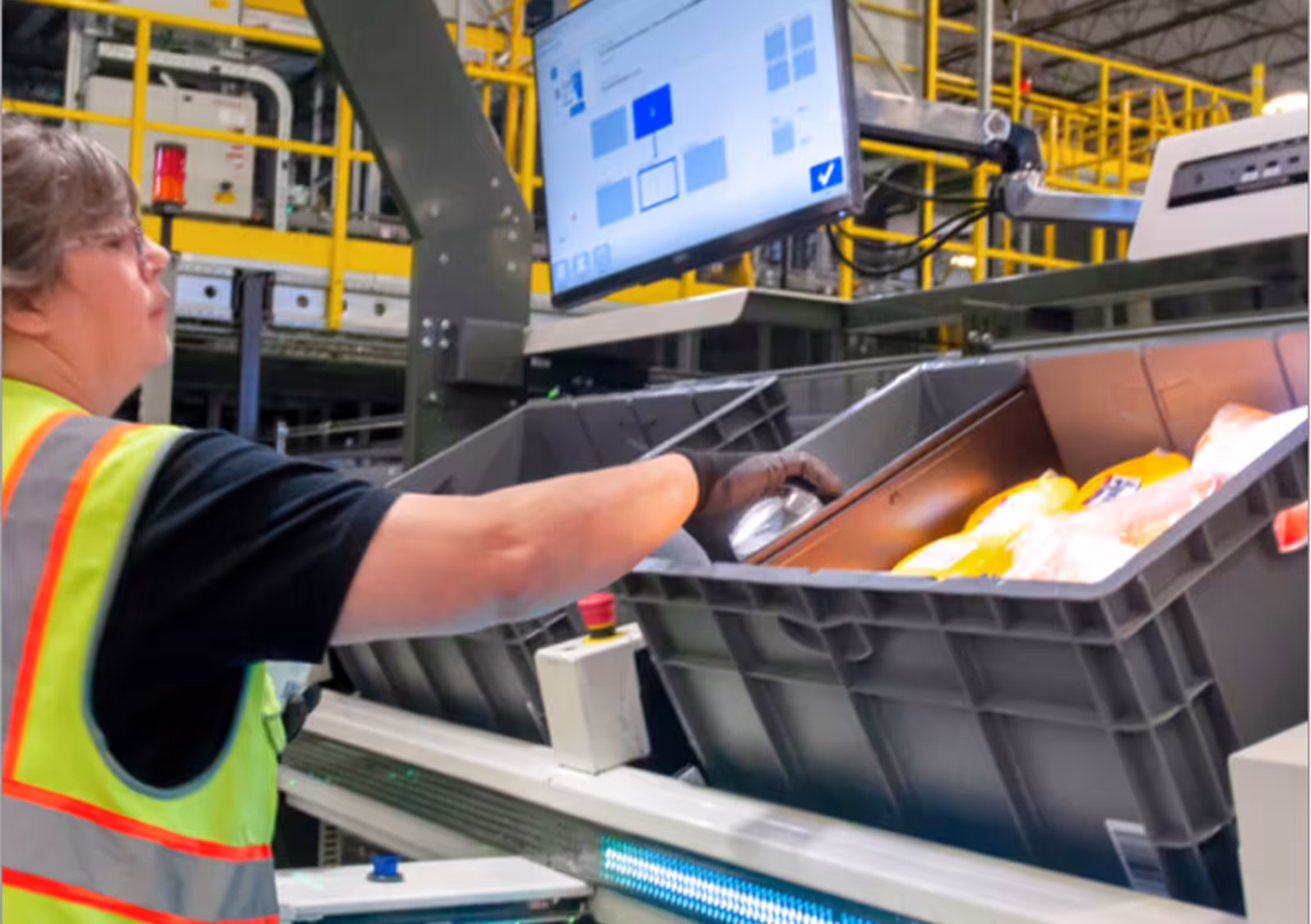 Warehouse worker using a screen-guided picking station, placing items into a plastic tote on an automated conveyor system.