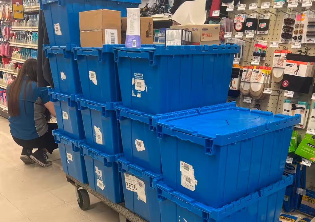 Store employee stocking shelves beside stacked blue plastic totes on a cart in a retail aisle.